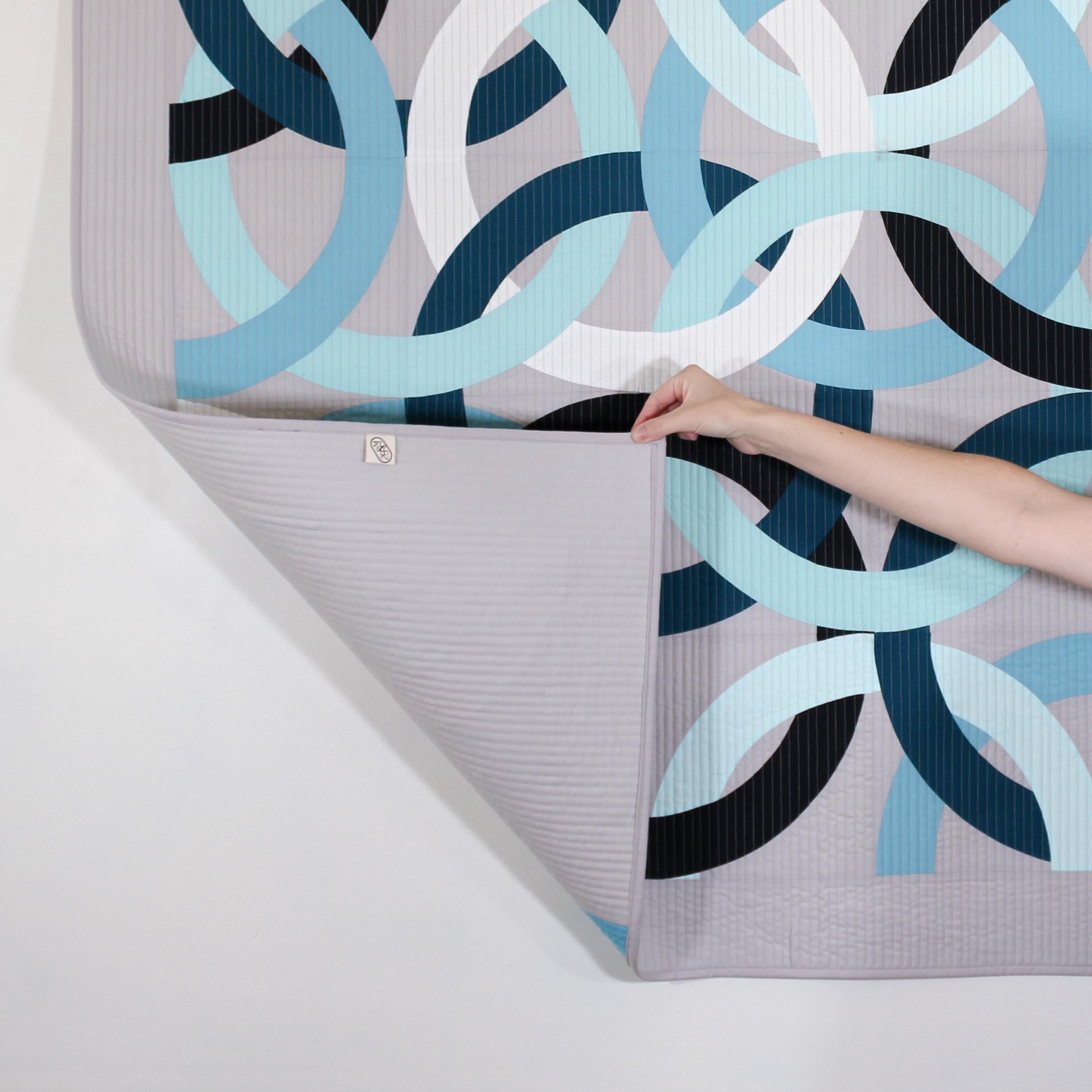 Geometric, modern, quilted wall hanging of interlocking rings in blue, black, and white on a gray background, with hand lifting up quilt corner to show backing of quilt