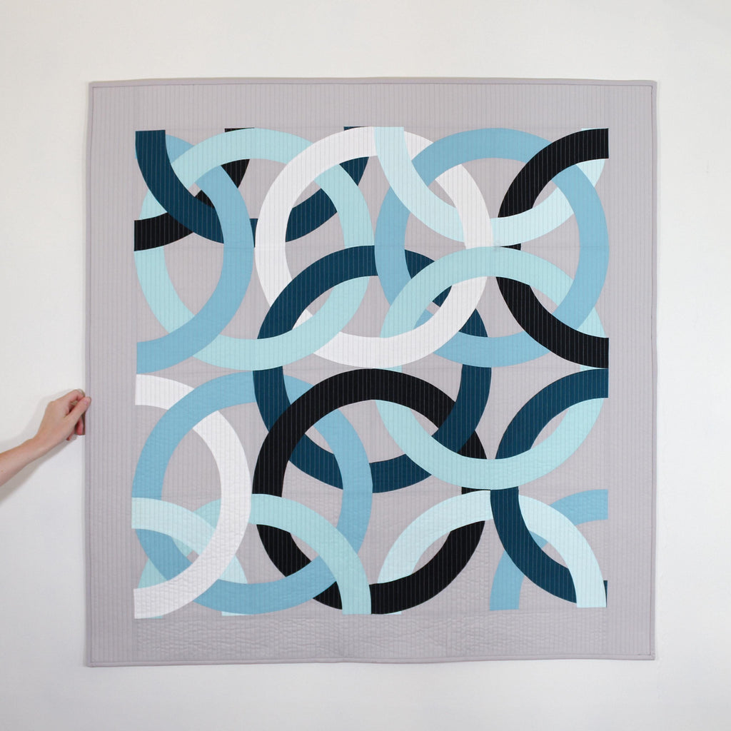 Geometric, modern, quilted wall hanging of interlocking rings in blue, black, and white on a gray background