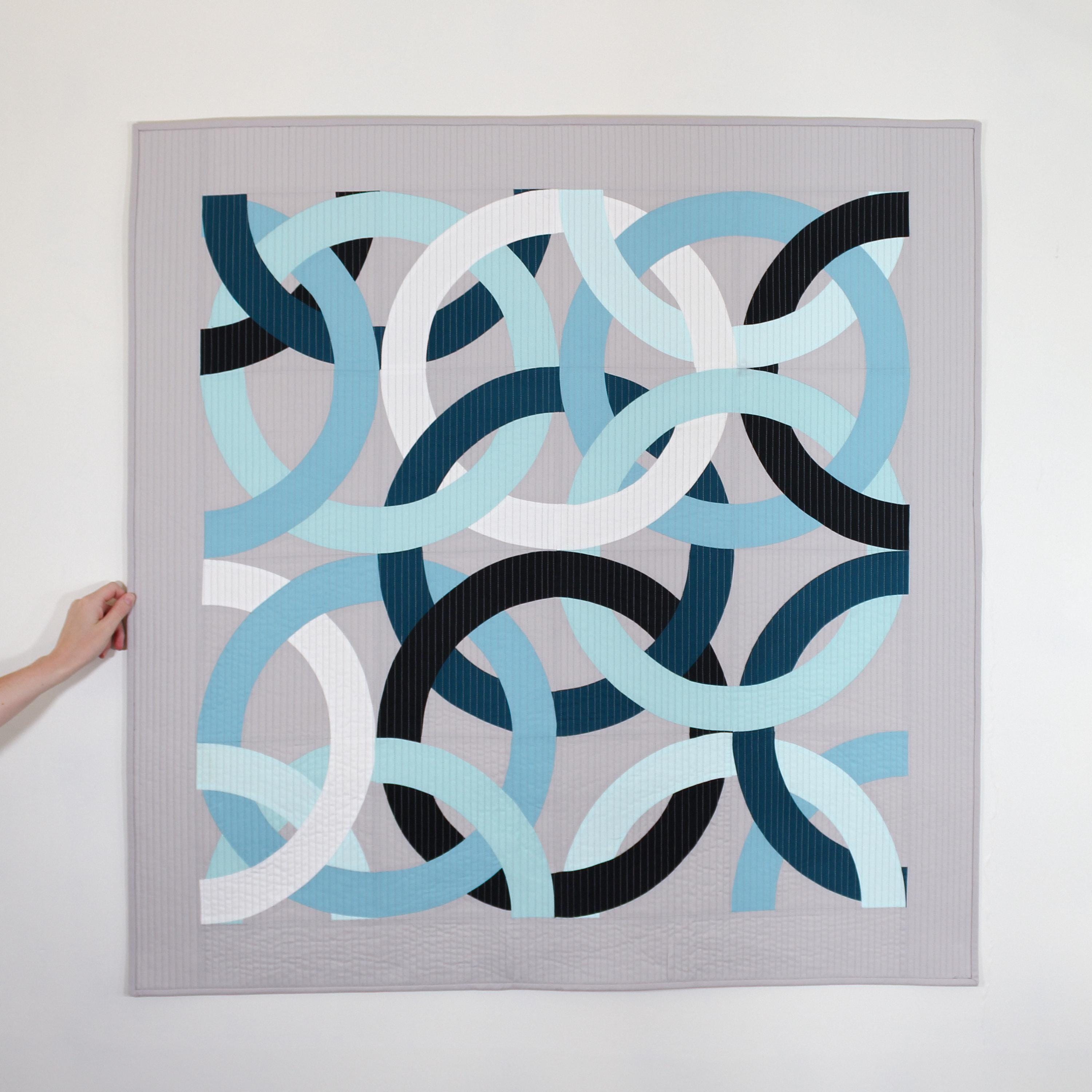 Geometric, modern, quilted wall hanging of interlocking rings in blue, black, and white on a gray background