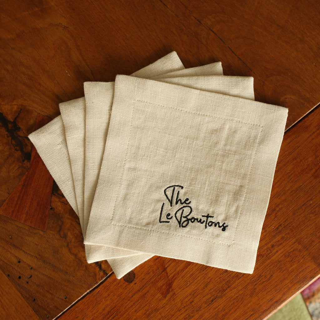 A set of four white linen cocktail napkins with custom black embroidered last name (The LeBoutons), stacked on a wooden tabletop