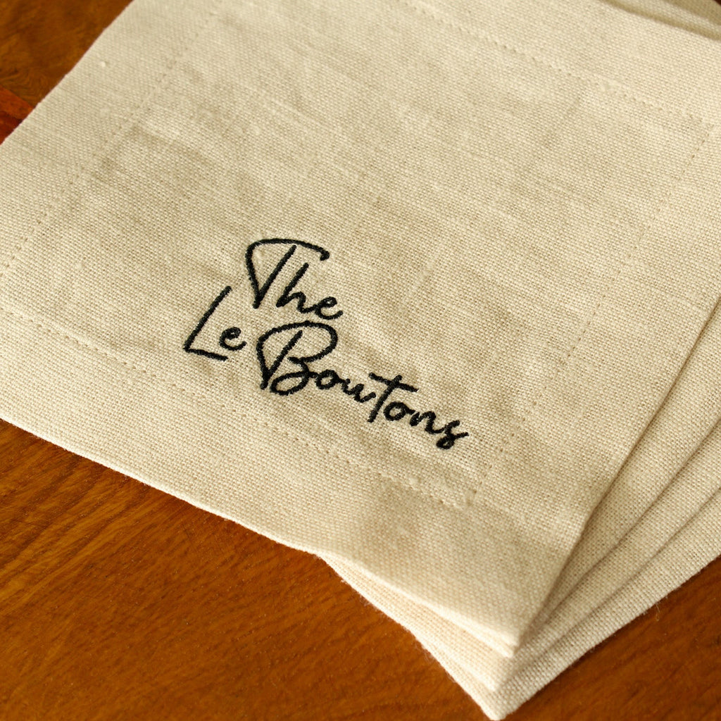 A set of four white linen cocktail napkins with custom black embroidered last name (The LeBoutons), stacked on a wooden tabletop