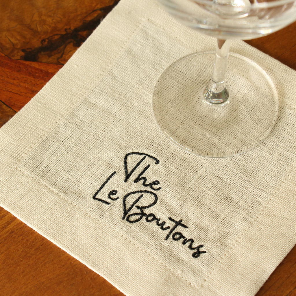 A white linen cocktail napkin with custom black embroidered last name (The LeBoutons) close up, with a wine glass on it