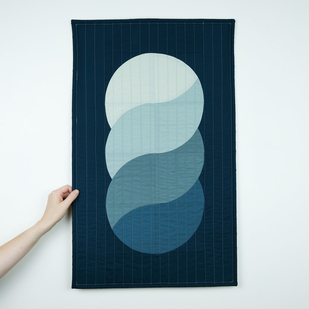 A hand holds a dark navy blue fabric wall hanging or tapestry against a plain white wall. The textile features a large central abstract wave-like design in stacked light blue and teal circles, with fine vertical stitched lines across the surface.