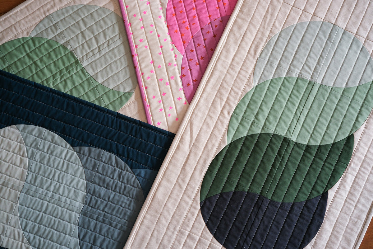 Close-up of quilted fabric panels with overlapping circular shapes in muted green, navy, pale blue, pink, and cream, arranged diagonally.