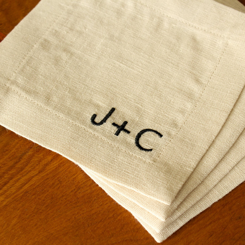 A set of four white linen cocktail napkins with custom black embroidered monograms, stacked on a wooden tabletop