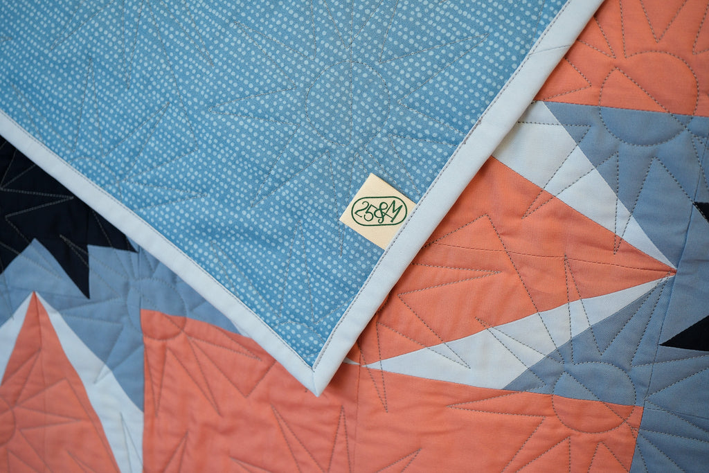 Close-up of a quilt with geometric patterns and a brand logo on a blue background.