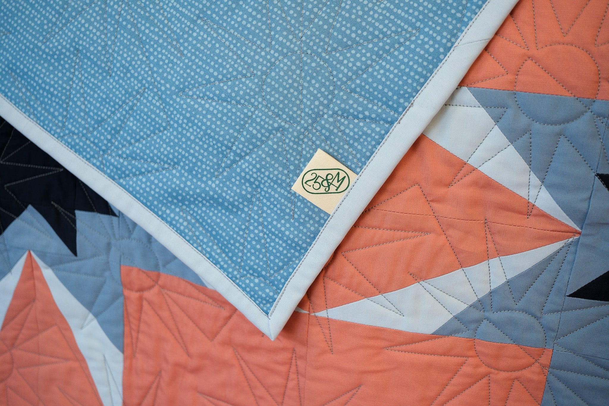 Close-up of a quilt with geometric patterns and a brand logo on a blue background.