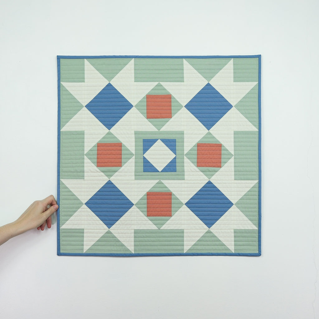 Hand holding a geometric quilt with blue, green, and red pattern on a white background