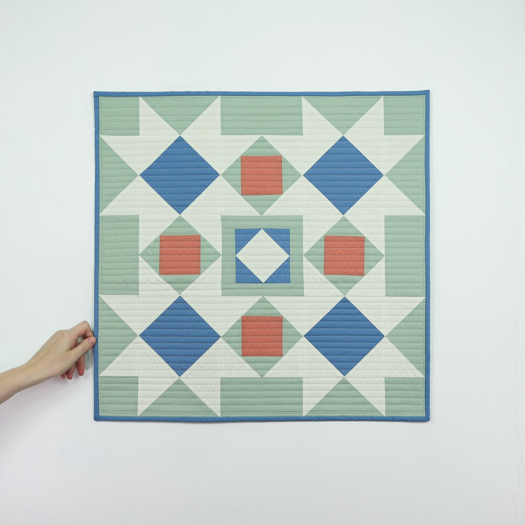 Hand holding a geometric quilt with blue, green, and red pattern on a white background