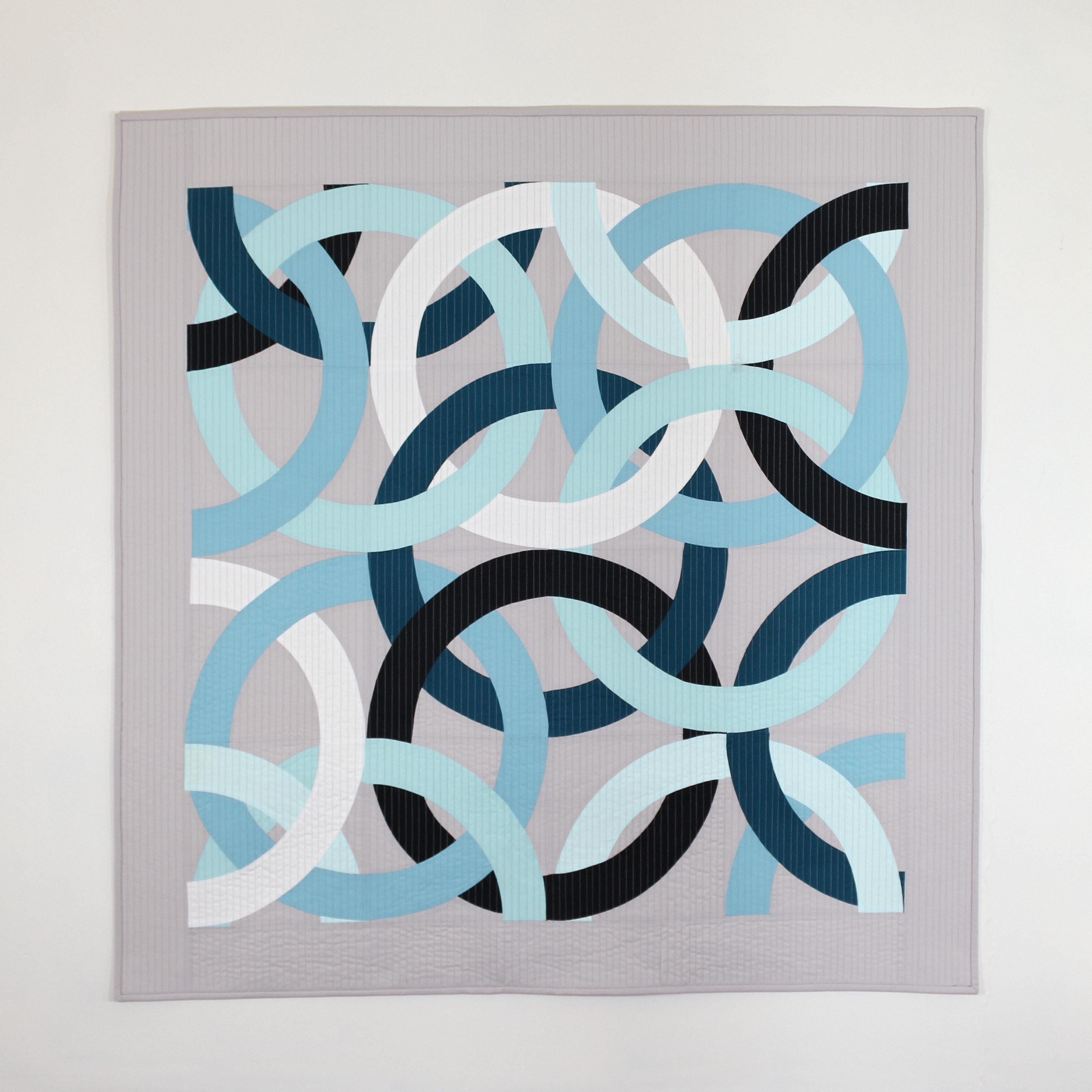 Geometric, modern, quilted wall hanging of interlocking rings in blue, black, and white on a gray background